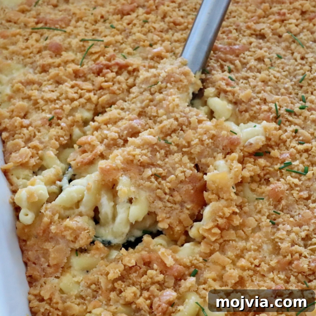Golden Cheez-It Mac and Cheese Bake 12 Close-up of creamy macaroni and cheese topped with golden, crispy Cheez-It cracker topping. A serving spoon is lifting up a portion, revealing gooey cheese beneath.