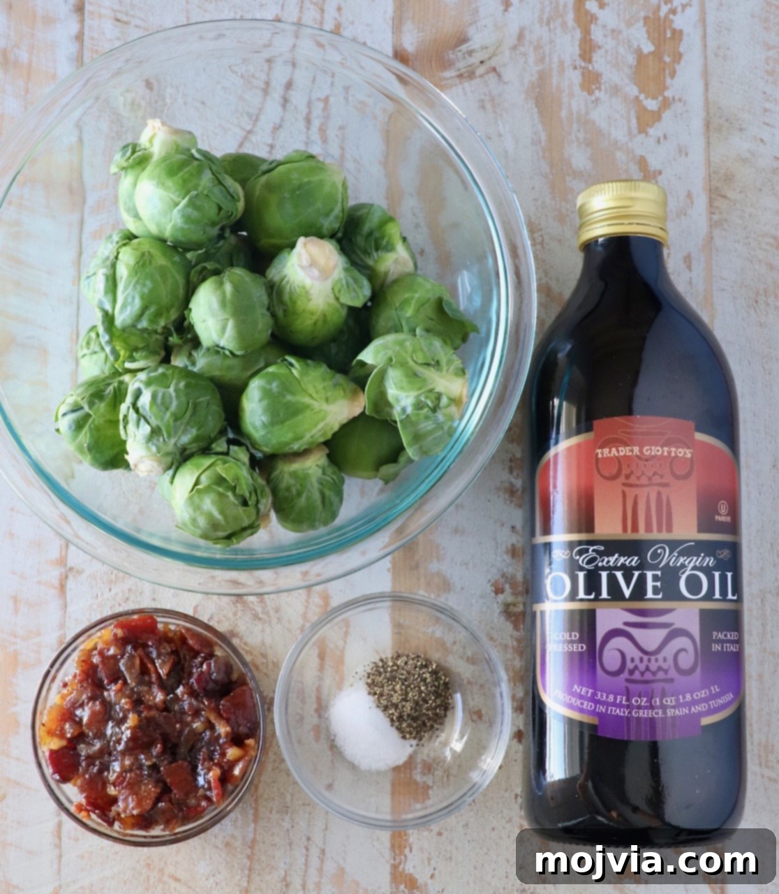 Sticky Bacon Brussels Sprouts 3 A bowl of Brussels sprouts, a bottle of olive oil, a small dish of bacon jam, and a bowl with salt and pepper are arranged on a wooden surface.