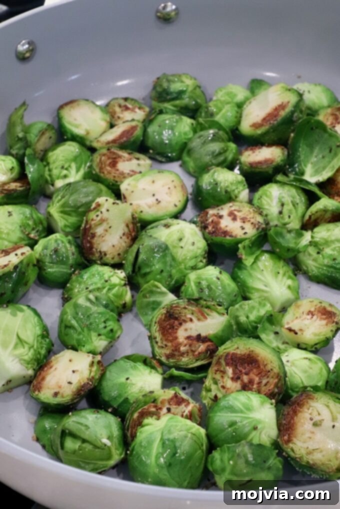 Sticky Bacon Brussels Sprouts 5 Brussels sprouts are halved and being sautéed in a pan. Their vibrant green color contrasts with golden-brown sear marks.