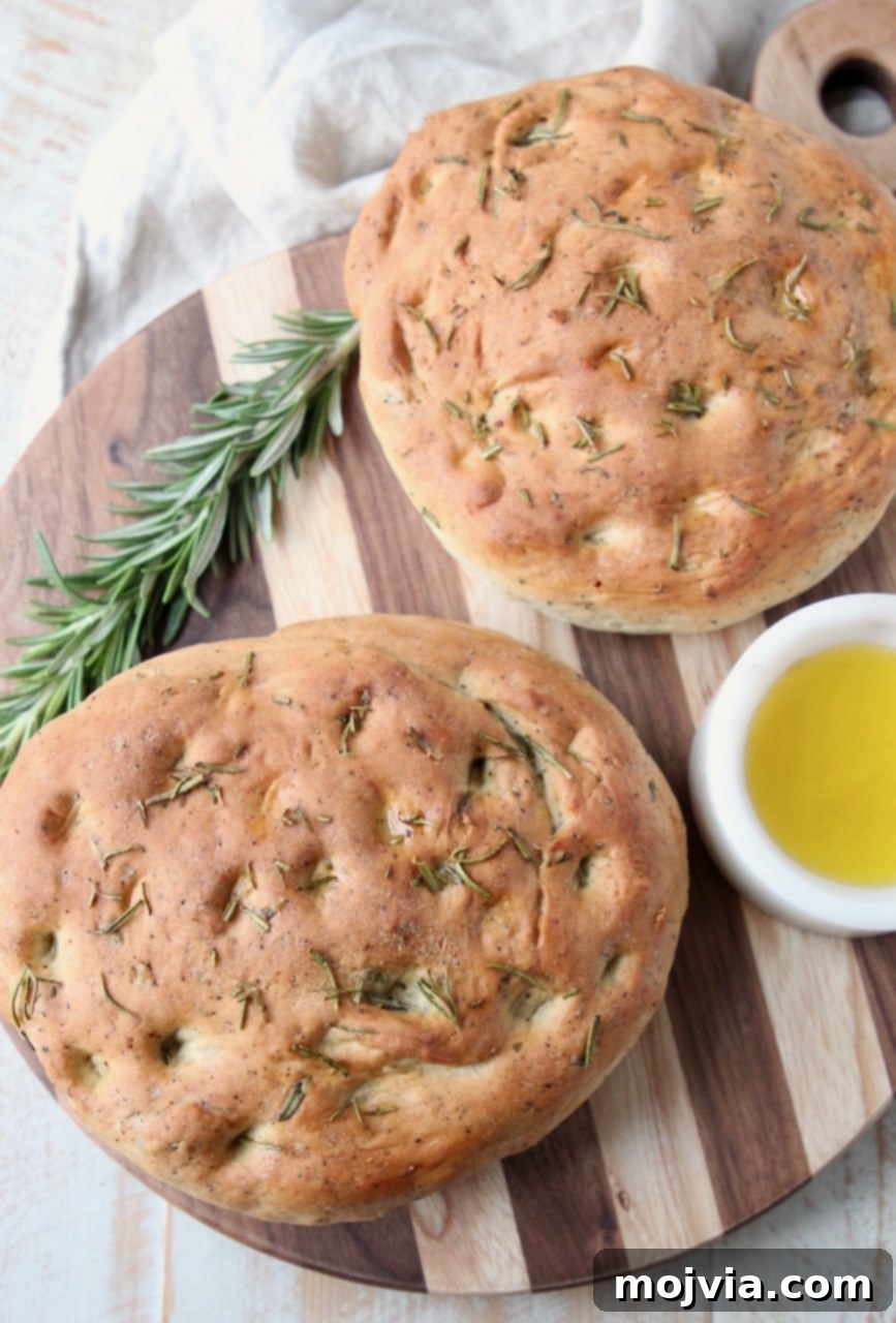 Fragrant Rosemary Focaccia 4 Two loaves of rosemary focaccia bread on wood cutting board with fresh sprig of rosemary and small white bowl of olive oil