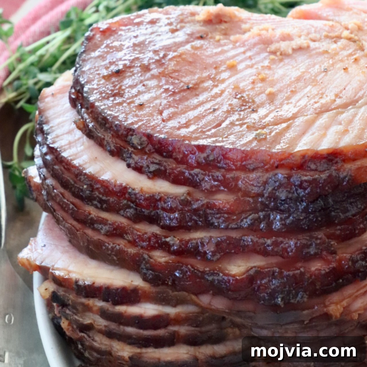 Sliced spiral ham glazed to a shiny brown, stacked on a white plate. Fresh herbs in the background and a red-striped cloth