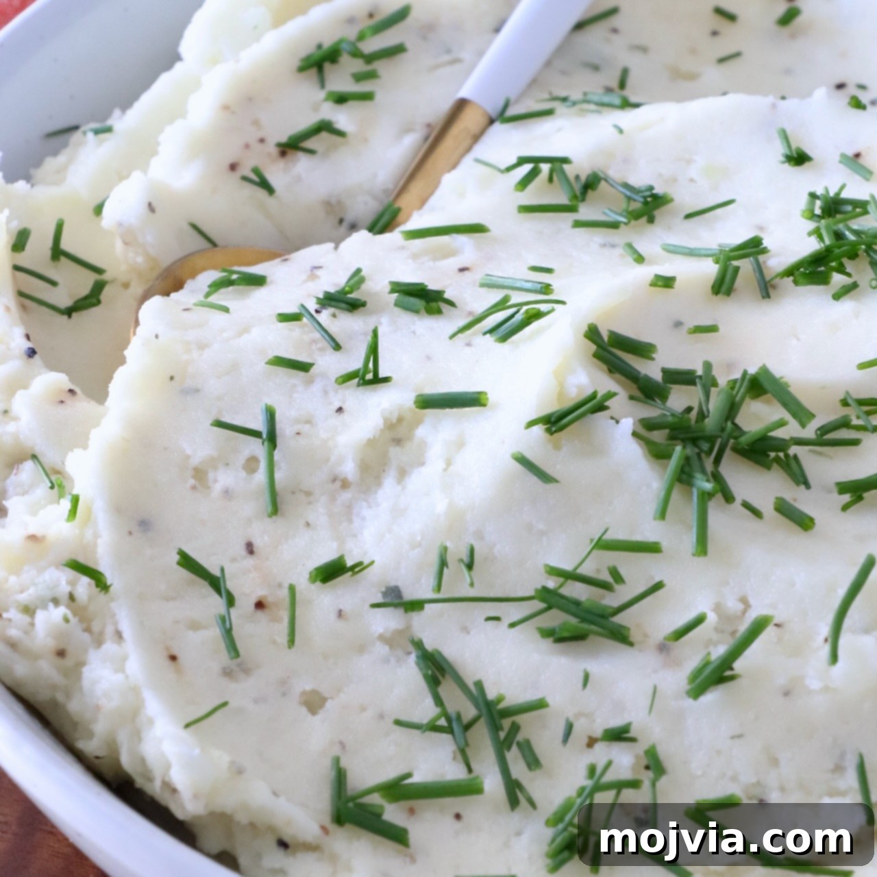 Creamy mashed potatoes garnished with fresh chopped chives in a white bowl. A gold and white spoon is dipped in the potatoes.