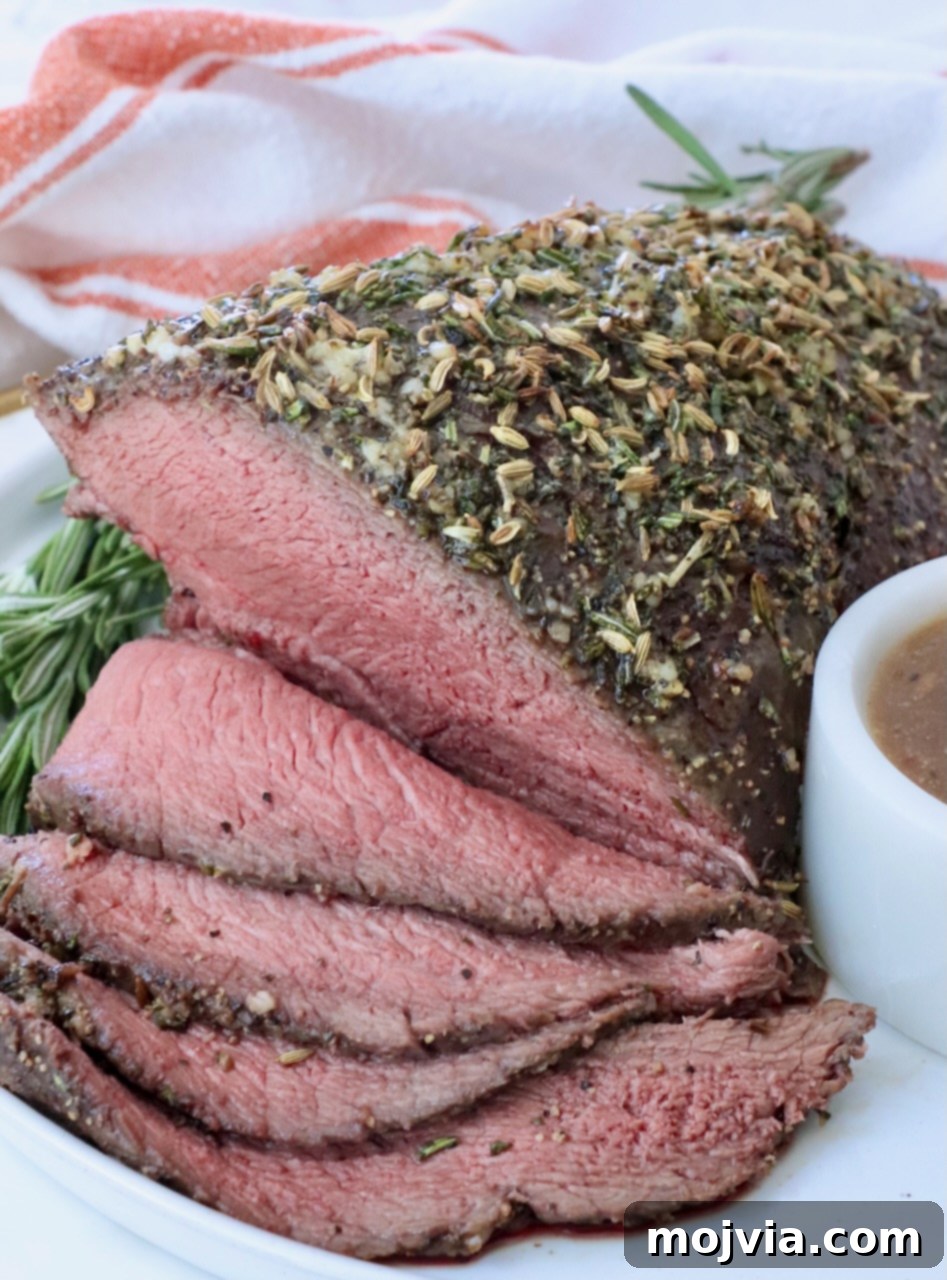 Tender Sirloin Tip Roast 2 Roast beef topped with herbs and spices, sliced to reveal a juicy pink interior. Garnished with rosemary and served on a white plate with gravy.
