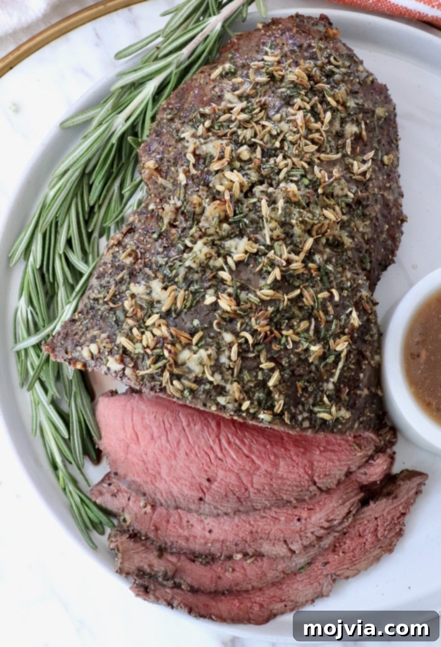 Tender Sirloin Tip Roast 9 Roasted beef topped with herbs, sliced on a white plate with a sprig of rosemary. A small bowl of gravy sits on the side.