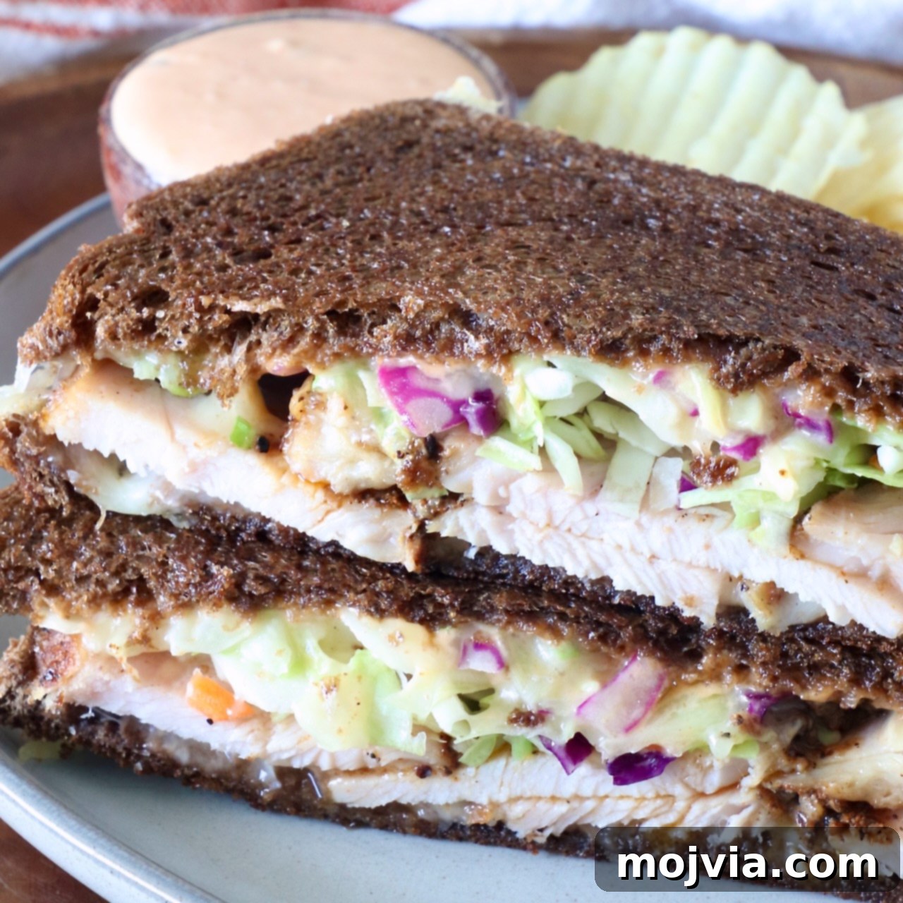 Golden Grilled Turkey Reuben 2 Close-up of a Rachel sandwich with dark rye bread filled with sliced turkey, creamy slaw, and sauce. Served on a plate with wavy potato chips and a dipping sauce.