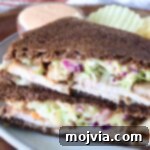 Close-up of a Rachel sandwich with dark rye bread filled with sliced turkey, creamy slaw, and sauce. Served on a plate with wavy potato chips and a dipping sauce.