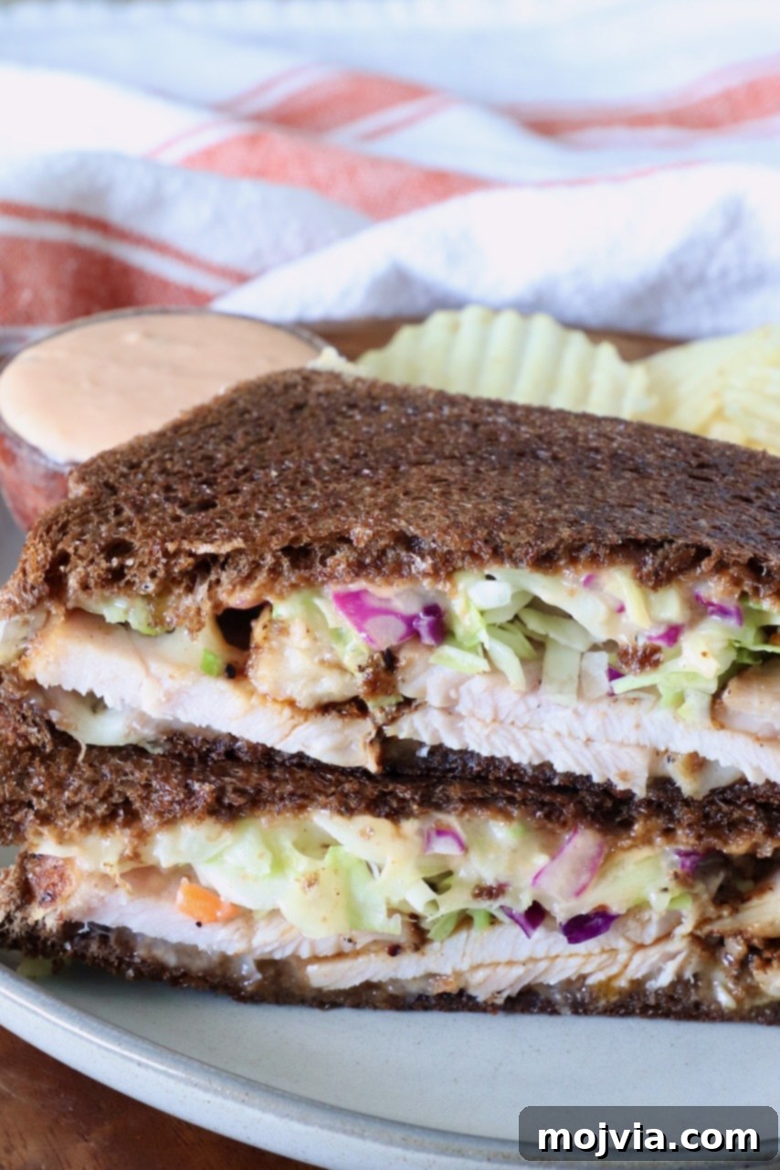 Golden Grilled Turkey Reuben 7 Close-up of a Rachel sandwich with dark rye bread filled with sliced turkey, creamy slaw, and sauce. Served on a plate with wavy potato chips and a dipping sauce.