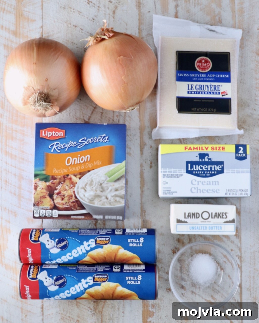 Savory French Onion Bites 3 Ingredients on a wooden surface include two yellow onions, a block of Gruyère cheese, a packet of onion soup mix, a block of cream cheese, tubes of crescent roll dough, a stick of butter, and a small dish of salt, all essential for creating French Onion Crescent Rolls.