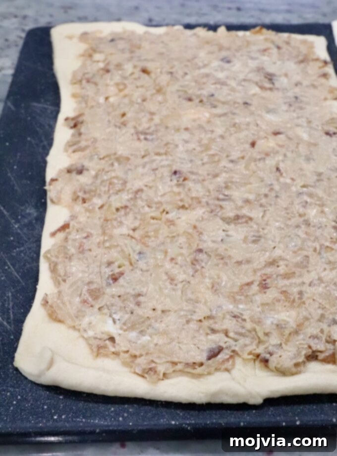 Savory French Onion Bites 6 Rectangular dough on a black cutting board, topped with a creamy mixture of caramelized onions and cheese, covering the entire surface, preparing for rolling.