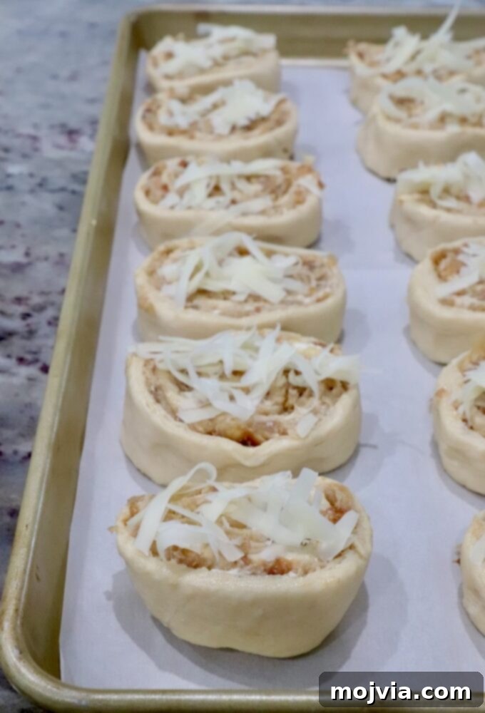 Savory French Onion Bites 8 Close-up of unbaked caramelized onion rolls on a parchment-lined baking sheet. Each roll is topped with shredded cheese, promising a golden, melty finish.