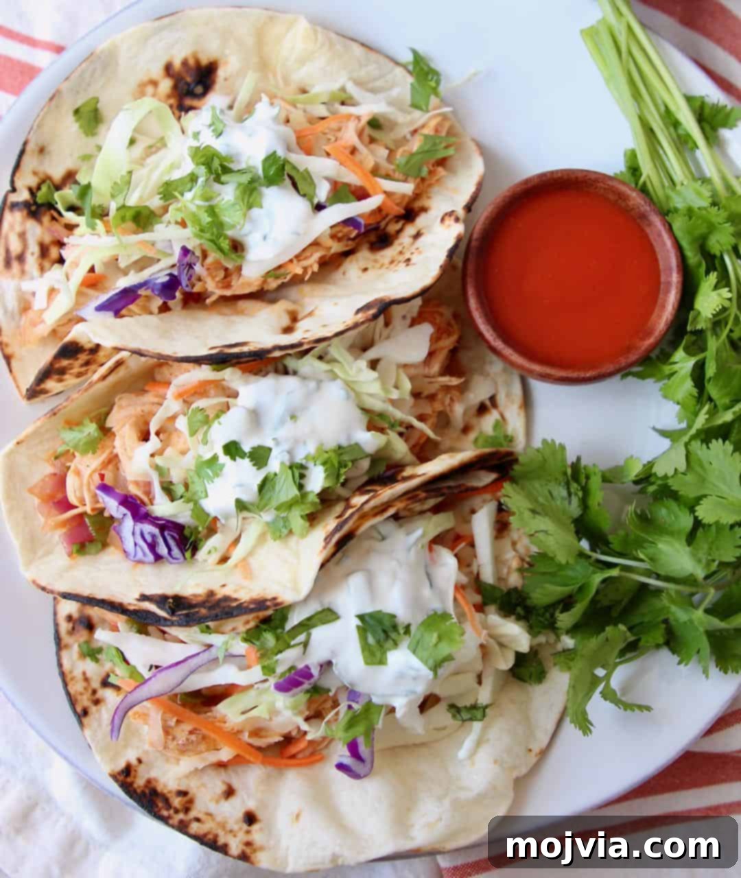Fiery Buffalo Chicken Tacos 2 Three buffalo chicken tacos on a white plate with a side of buffalo sauce, garnished with fresh herbs and lime wedges.