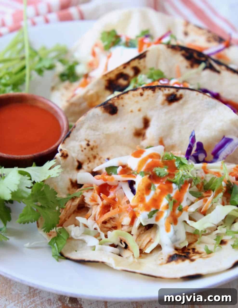 Fiery Buffalo Chicken Tacos 3 Two buffalo chicken tacos with a fresh coleslaw and a small bowl of buffalo sauce on a pristine white plate.