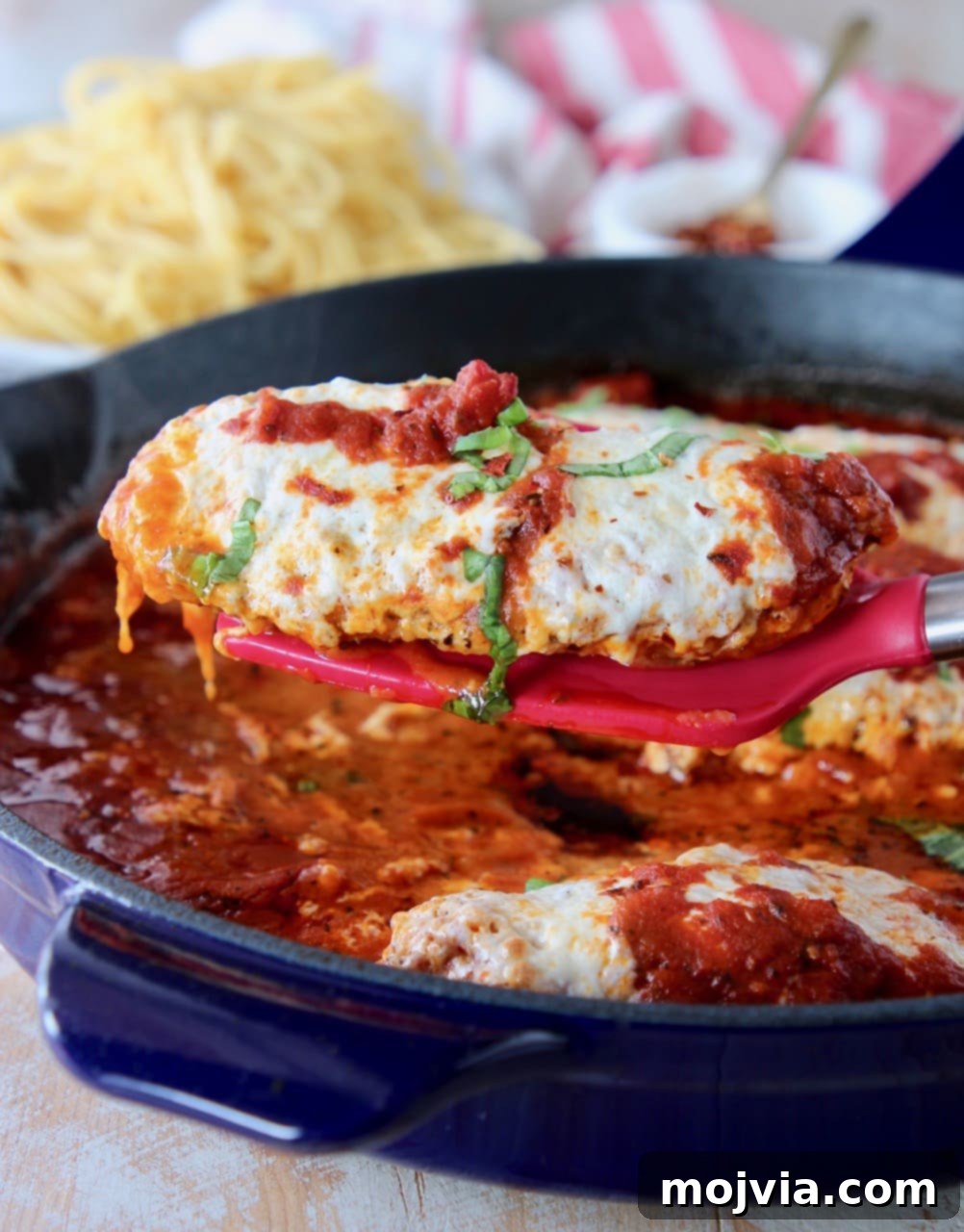 Red spatula holding a piece of chicken parmesan out of a large blue skillet, garnished with fresh basil.