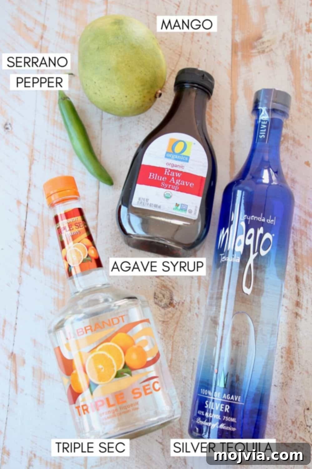 Serrano Mango Margarita Heatwave 3 A flat lay photograph showcasing all the fresh ingredients for spicy mango margaritas: silver tequila, triple sec, agave nectar, a ripe mango, fresh lime, and serrano chili peppers.
