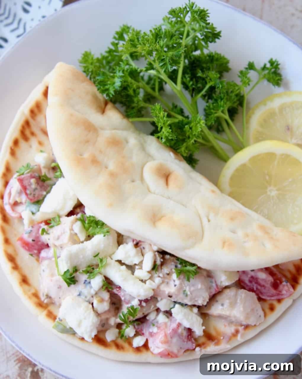Zesty Greek Chicken Pita Pocket 4 An inviting overhead shot of Greek Chicken Salad served in two halves of warm pita bread, garnished with fresh parsley and lemon wedges, on a rustic white plate.