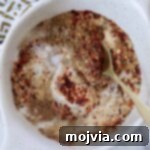 A white bowl contains a mix of red chili flakes, brown spices, and white salt, stirred with a golden spoon. A patterned cloth is partially visible.