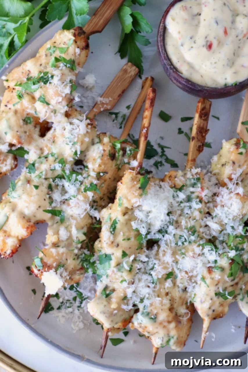Garlic Parmesan Chicken Kebabs 2 Garlic Parmesan Chicken Skewers: Cooked chicken skewers topped with creamy sauce and shredded cheese, garnished with chopped parsley, served beside a small bowl of dipping sauce.