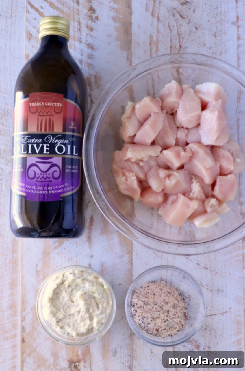 Garlic Parmesan Chicken Kebabs 3 Ingredients for Garlic Parmesan Chicken Skewers: Olive oil next to a bowl of raw chicken chunks, with small bowls of creamy sauce and seasoning on a rustic wooden surface.
