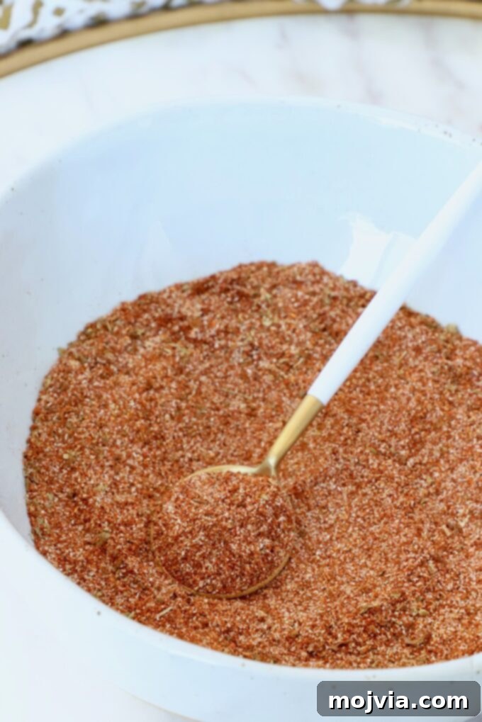 Chorizo Spice Blend 4 Close-up of a white bowl filled with reddish-brown spice mix, with a gold and white spoon resting on top.