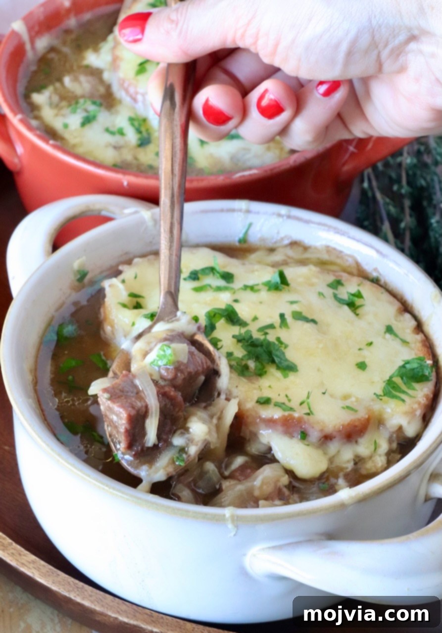 Caramelized Onion Beef Braise 2 A bowl of French onion beef stew topped with melted cheese and herbs. A hand with red nail polish holds a spoon, lifting a piece of beef and onion out of the bowl.