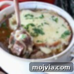 A bowl of French onion beef stew topped with melted cheese and herbs. A hand with red nail polish holds a spoon, lifting a piece of beef and onion out of the bowl.