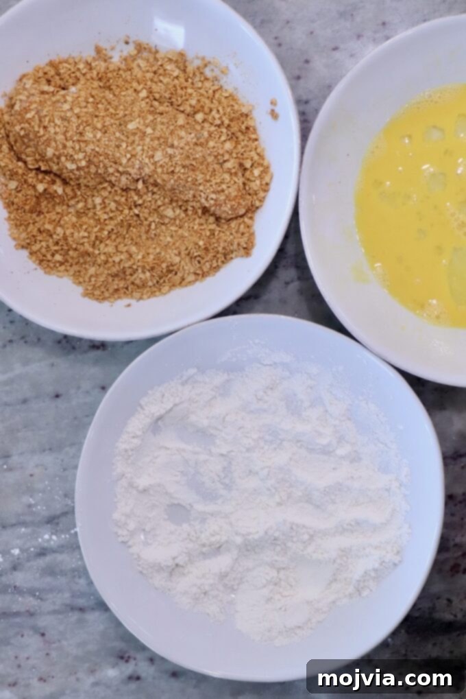 Fiesta Chicken Parmesan 4 Three bowls on a marble surface contain ingredients for breading: flour, beaten eggs, and tortilla chip crumbs. The setup is ready for coating food.