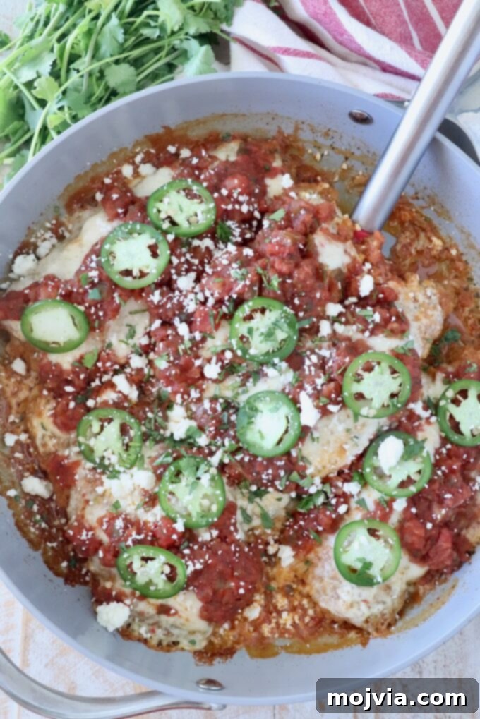 Fiesta Chicken Parmesan 9 A skillet filled with chicken topped with vibrant red salsa, green jalapeño slices, and crumbled cotija cheese. Fresh cilantro and a striped towel are nearby.