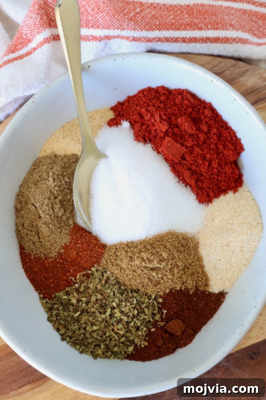 Mexican Street Taco Al Pastor Blend 2 A white bowl filled with colorful spices, including red chili powder, white salt, brown cumin, and green oregano. A gold spoon rests on the side.