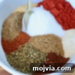 A white bowl filled with colorful spices, including red chili powder, white salt, brown cumin, and green oregano. A gold spoon rests on the side.