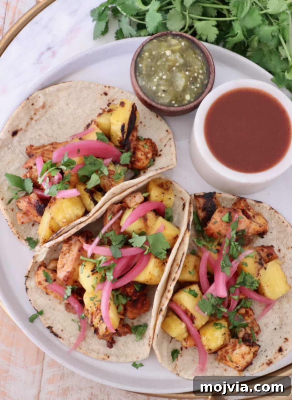 Vibrant Al Pastor Chicken 6 Three delicious tacos filled with grilled chicken, pineapple, pickled onions, and cilantro, served on a plate next to small bowls of green salsa and hot sauce.
