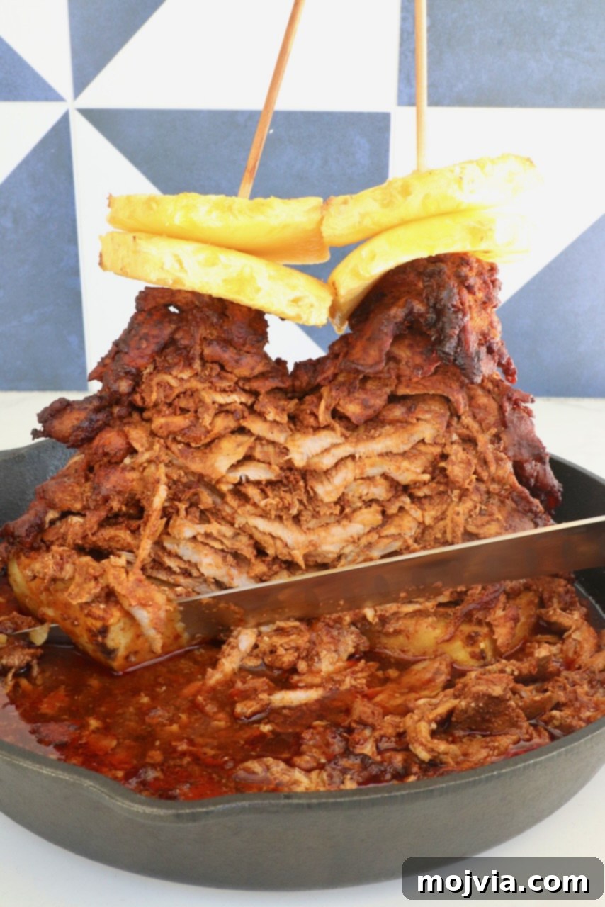 Sweet & Spicy Al Pastor Pork 2 A stack of marinated, cooked pork layered on skewers, topped with pineapple slices, in a cast iron skillet. A knife slices through the juicy, cooked meat.