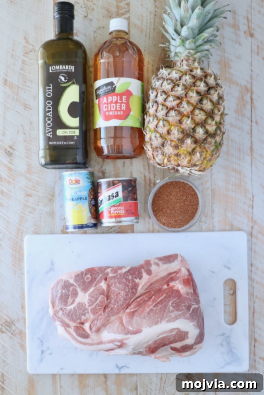 Sweet & Spicy Al Pastor Pork 3 Ingredients for al pastor are arranged on a wooden surface. Items include avocado oil, apple cider vinegar, a whole pineapple, canned pineapple juice, chipotle peppers, a bowl of spices, and a raw pork shoulder on a marble board.