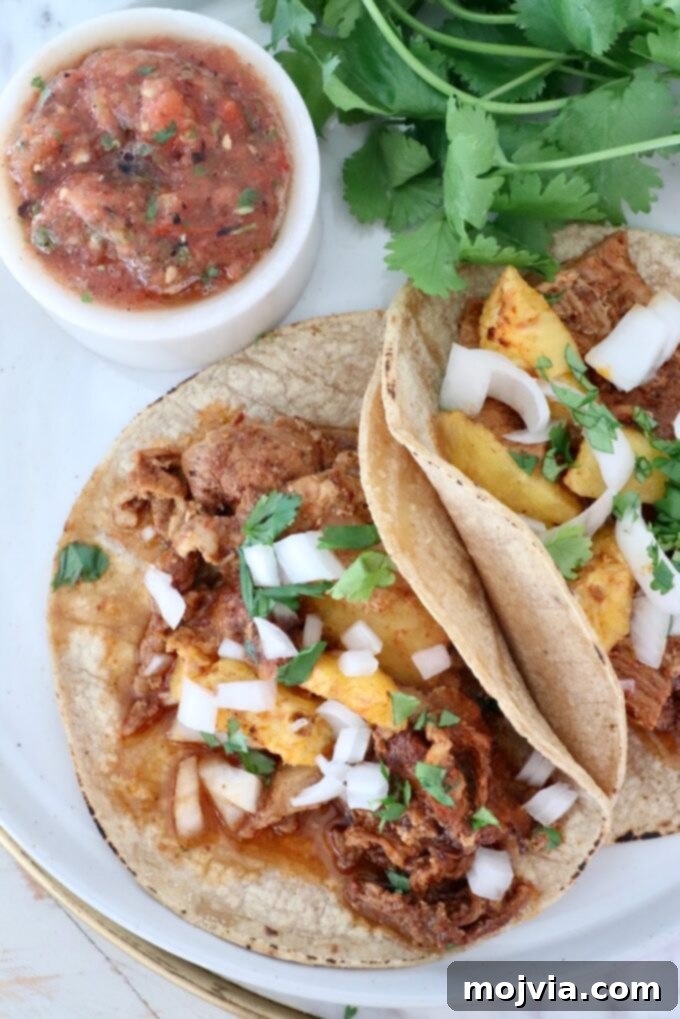 Sweet & Spicy Al Pastor Pork 7 Two tacos with pork al pastor, pineapple, onion, and cilantro on tortillas, accompanied by a bowl of salsa and fresh cilantro, arranged on a plate.