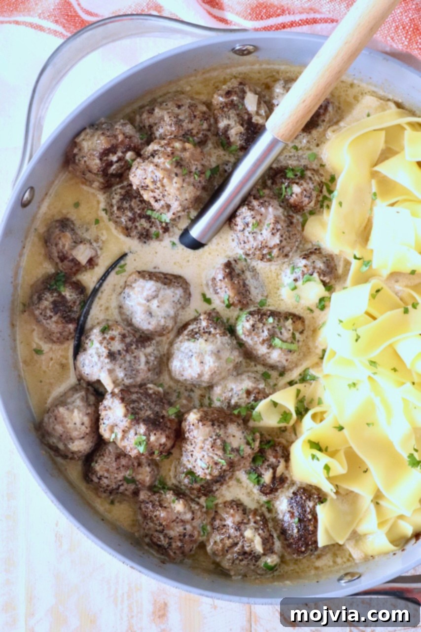 Creamy Peppercorn Meatballs 2 A pan filled with meatballs au poivre in a creamy sauce garnished with fresh herbs, alongside wide egg noodles. A ladle rests nearby.