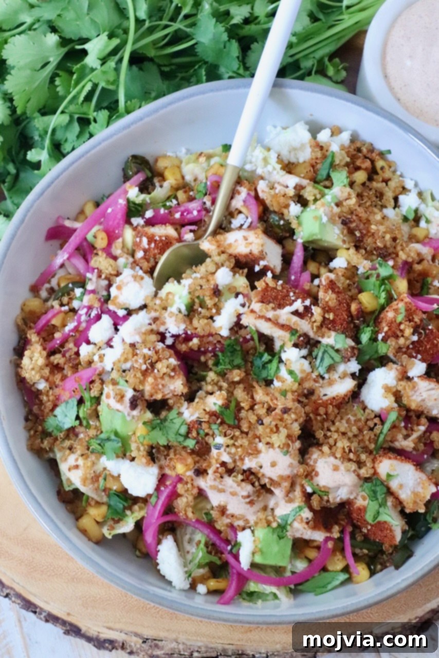 A vibrant salad in a bowl featuring quinoa, grilled chicken, avocado, corn, pickled onions, and crumbled cheese. Fresh cilantro and dressing are next to it.