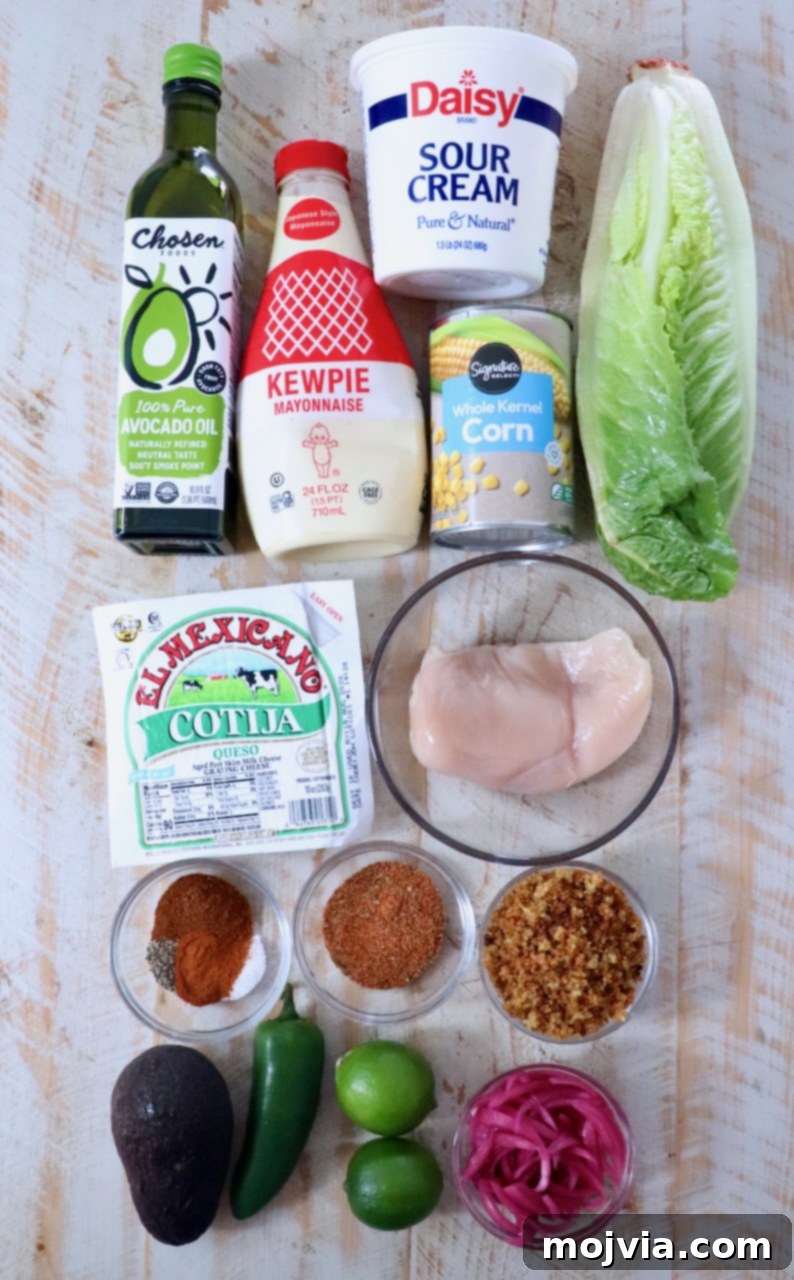 Ingredients on a wooden surface include avocado oil, sour cream, mayonnaise, corn, lettuce, chicken breast, cotija cheese, spices, avocado, jalapeño, limes, and pickled onions.