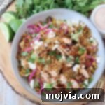 A vibrant salad in a bowl featuring quinoa, grilled chicken, avocado, corn, pickled onions, and crumbled cheese. Fresh cilantro and dressing are next to it.
