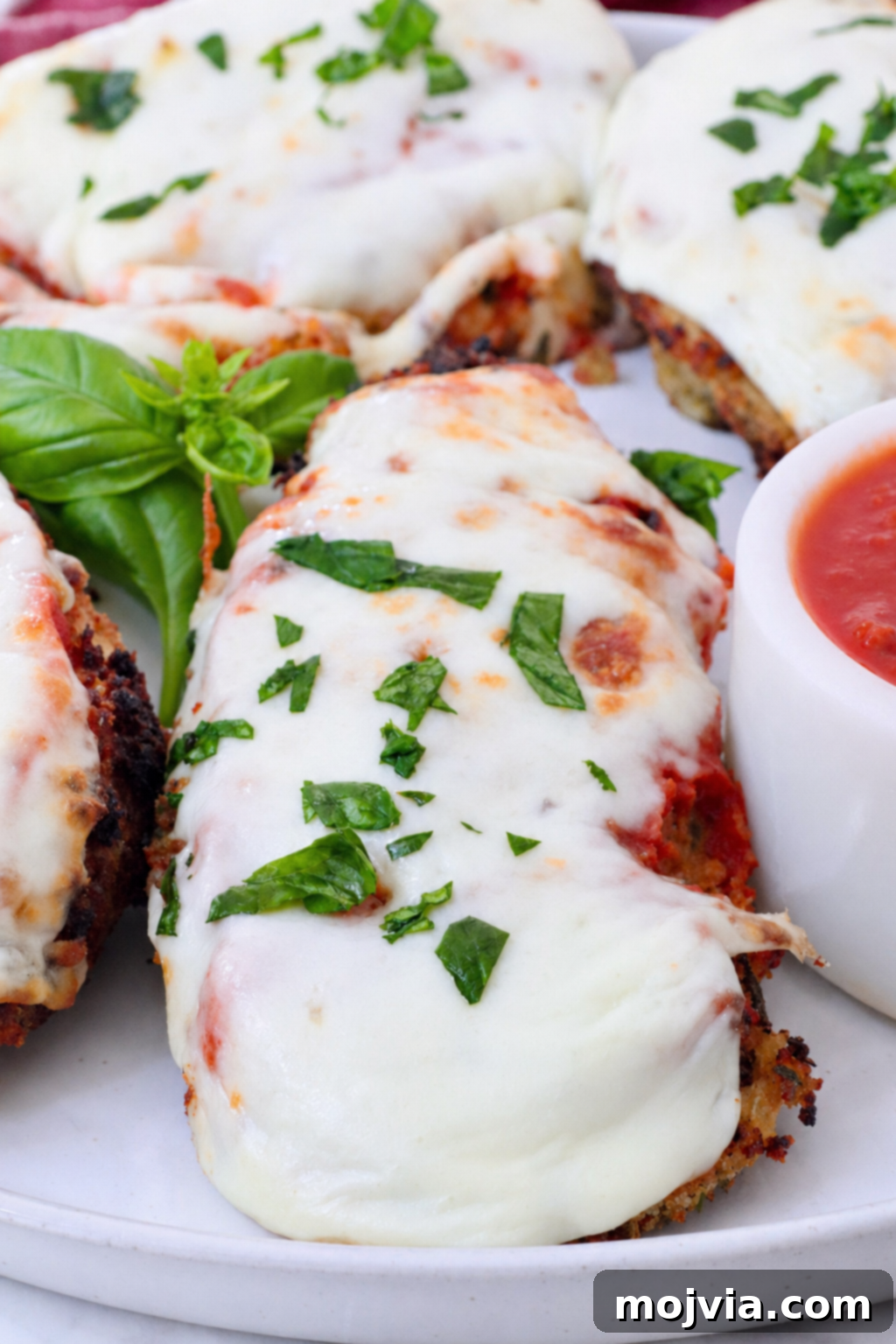 The Ultimate Air Fryer Chicken Parmesan 2 Crispy pieces of Air Fryer Chicken Parmesan on a plate with fresh basil and a small bowl of marinara sauce on the side, ready to be served.