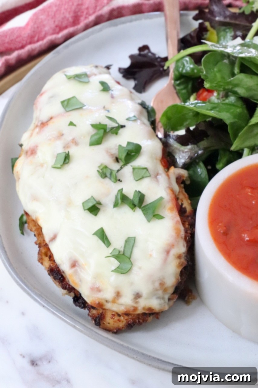 The Ultimate Air Fryer Chicken Parmesan 12 A perfectly cooked piece of Air Fryer Chicken Parmesan on a white plate, accompanied by a fresh side salad, a fork, and a small bowl of marinara sauce, ready to be enjoyed.
