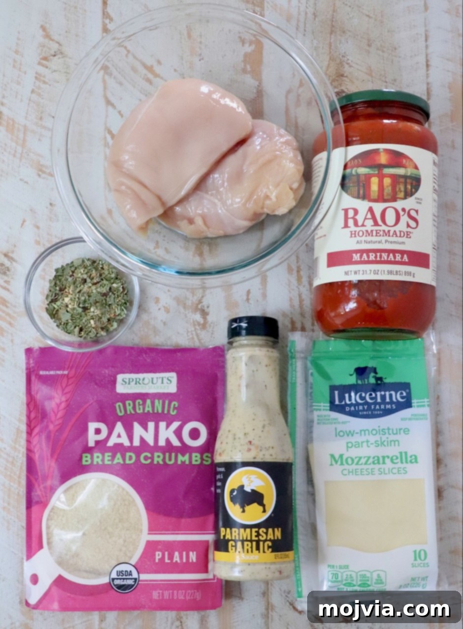 The Ultimate Air Fryer Chicken Parmesan 5 Ingredients for Air Fryer Chicken Parmesan laid out on a wooden surface: raw chicken breasts in a bowl, a jar of marinara sauce, a bag of panko bread crumbs, a bottle of Parmesan garlic dressing, slices of mozzarella cheese, and a bowl of fresh herbs.