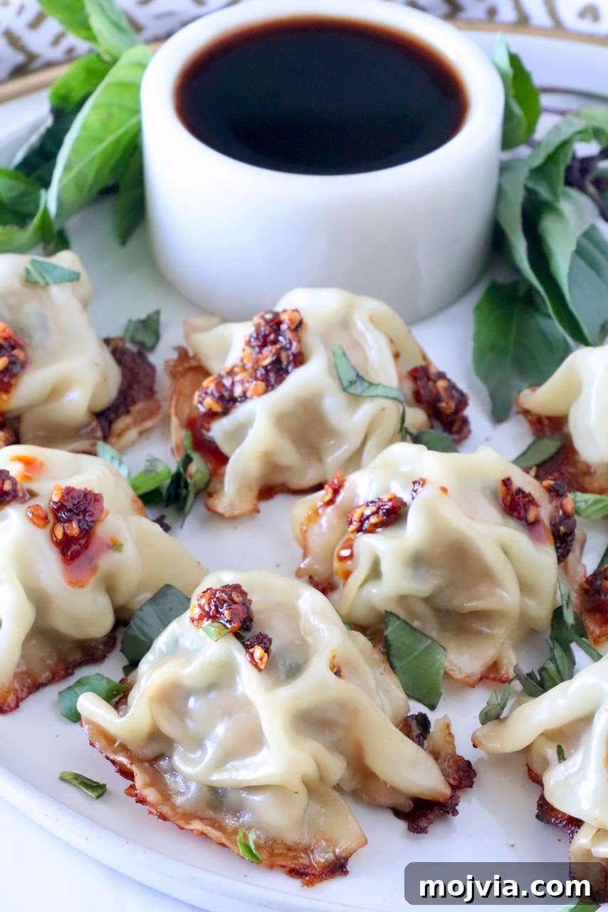 Thai Basil Chicken Dumplings: Flavorful Asian-Style Potstickers 2 Thai basil chicken dumplings on a plate with a small bowl of dark sauce behind the dumplings and fresh Thai basil leaves on the side