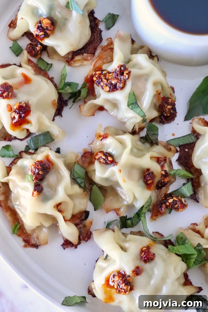 Thai Basil Chicken Dumplings: Flavorful Asian-Style Potstickers 12 cooked blanket dumplings on a plate topped with thai basil and chili crisp, with a small bowl of dipping sauce on the side