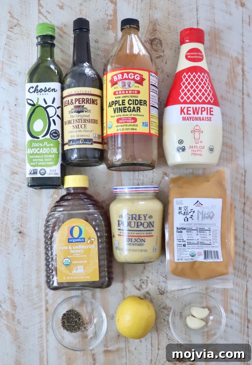 Miso Caesar Dressing Recipe: Creamy Umami Salad Dressing 5 A variety of ingredients are arranged on a wooden surface, including avocado oil, Worcestershire sauce, apple cider vinegar, Kewpie mayonnaise, honey, Dijon mustard, miso paste, a lemon, garlic cloves, and a small bowl of pepper.