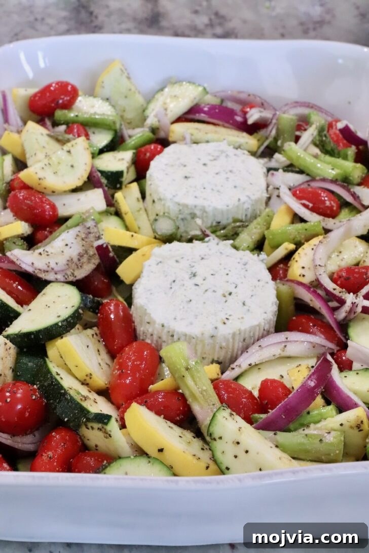 Creamy Boursin Pasta Primavera with Spring Vegetables 7 A white dish filled with vibrant chopped vegetables, including cherry tomatoes, zucchini, squash, and red onion, surrounds two seasoned cheese rounds.