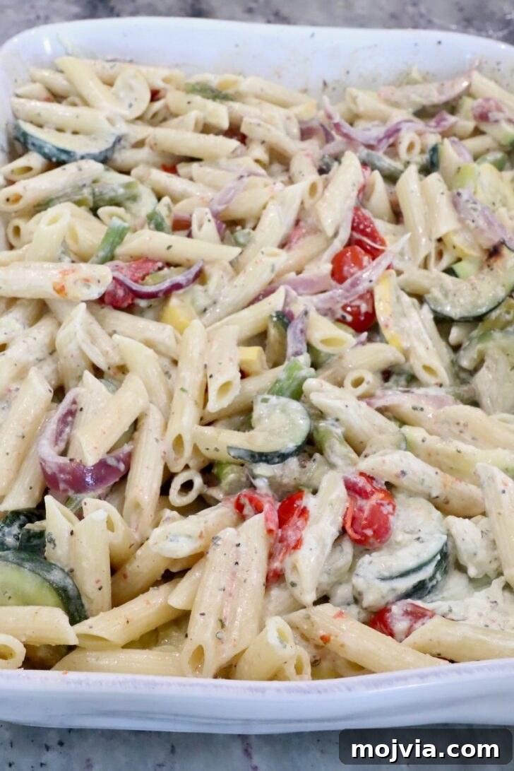 Creamy Boursin Pasta Primavera with Spring Vegetables 9 Creamy pasta bake with penne, zucchini, cherry tomatoes, red onions, and asparagus in a white casserole dish.
