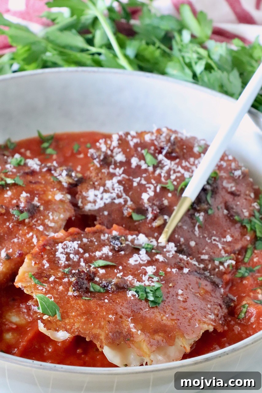 chicken ravioli with a mozzarella cheese crust in a bowl of marinara sauce with a fork, topped with fresh parsley and parmesan cheese