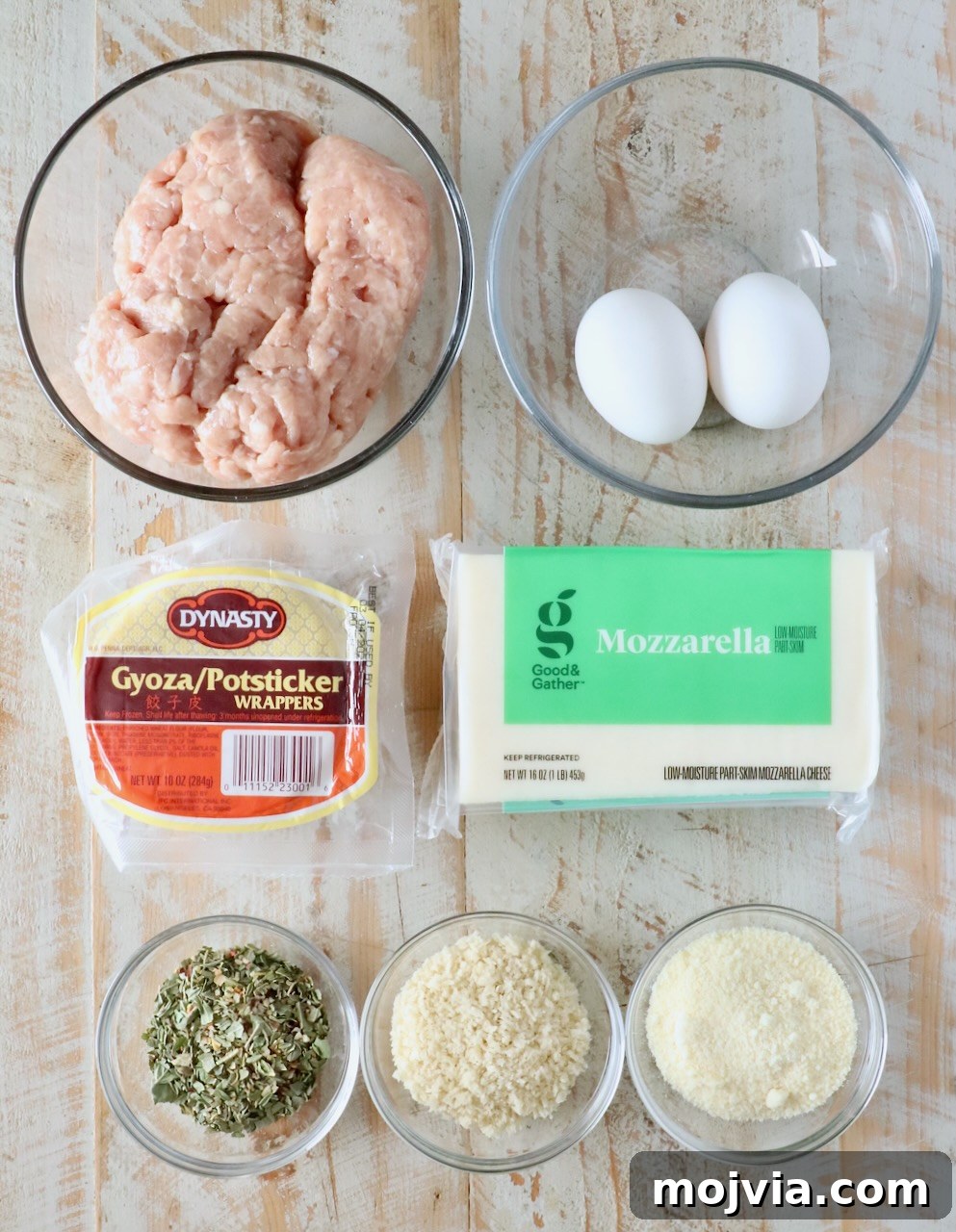 A top-down view of cooking ingredients on a wooden surface: ground chicken, two eggs, gyoza wrappers, mozzarella cheese, herbs, breadcrumbs, and grated cheese in bowls.