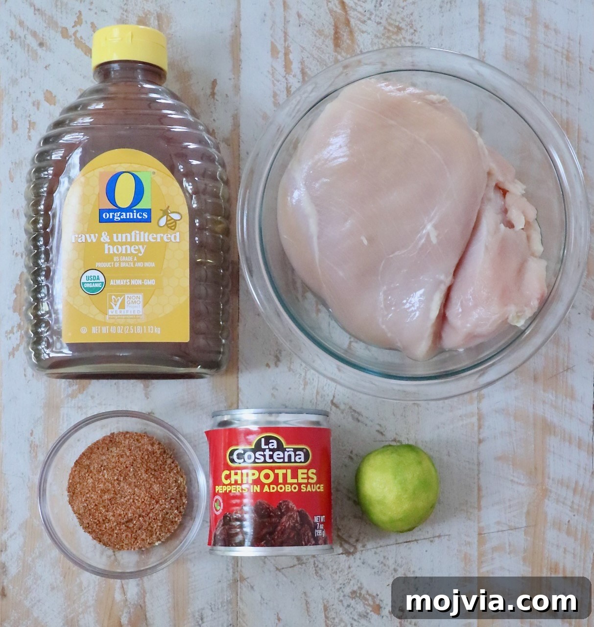 Chipotle Honey Chicken Tacos Made in the Air Fryer 5 Ingredients for cooking are arranged on a wooden surface: raw honey in a bottle, raw chicken breasts in a bowl, chipotle peppers in a can, a lime, and a bowl of taco seasoning.