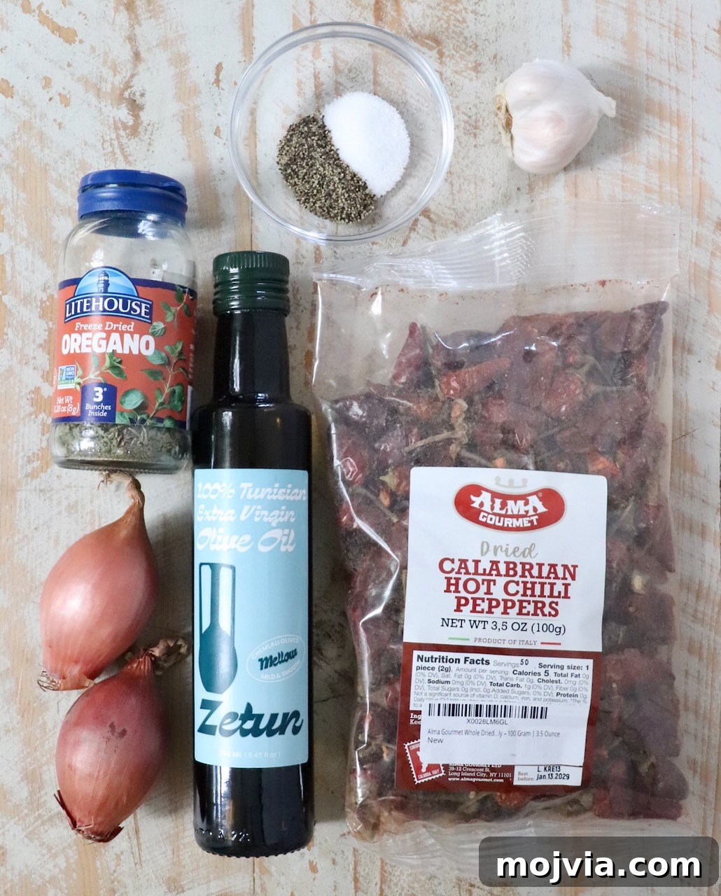 Calabrian Chili Crisp: Spicy Oil Recipe & Flavor Uses 5 Ingredients on a rustic wooden surface include a bottle of olive oil, a jar of dried oregano, dried Calabrian chili peppers, two shallots, a garlic bulb, and a bowl of salt and pepper.