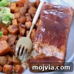 Grilled Salmon with Maple-Glazed Sweet Potatoes 9 A plate of glazed salmon fillet, accompanied by seasoned sweet potato cubes and a side salad. A gold fork rests on the plate.